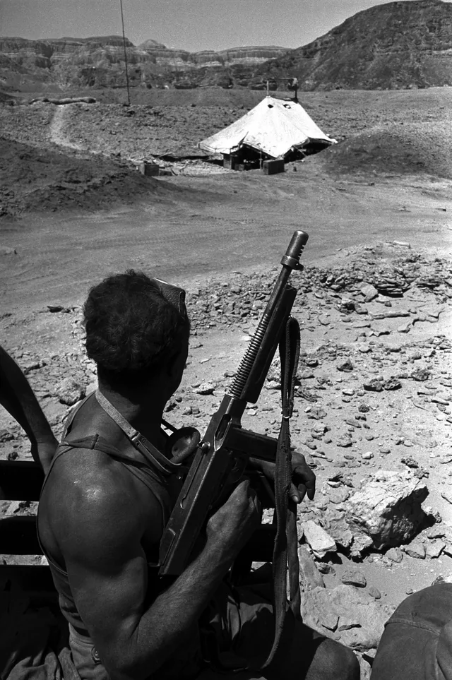 Vertical black-and-white photograph of a back-facing armed figure and rifle overlooking a canvas shelter in a rocky desert valley.