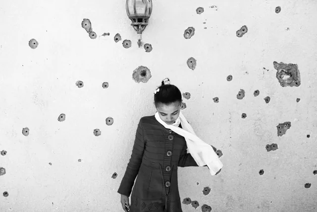 Black-and-white photograph of an apparently young person standing with head lowered before a white wall marked by many dark spots, beneath a caged lamp.