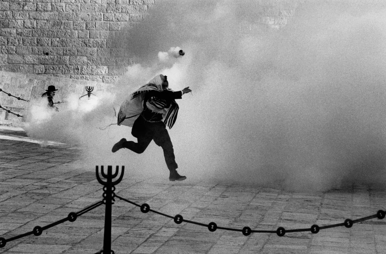 Black-and-white photograph of a person running through smoke at the Western Wall, with a canister in the air and a metal menorah barrier in the foreground.