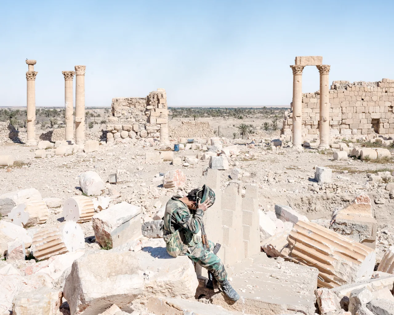 Color photograph of a soldier holding a helmet to his face while seated among pale rubble and standing columns at Palmyra under a clear blue sky.